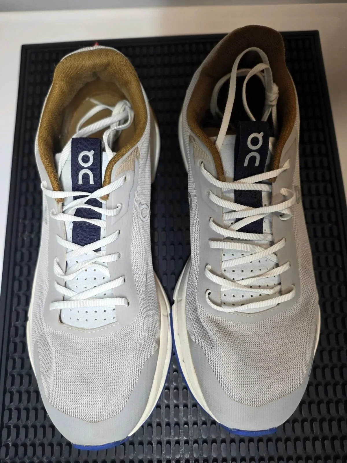 Pair of white and beige athletic sneakers with white laces, black and blue logo on tongue, and brown interior, placed on a black textured surface.