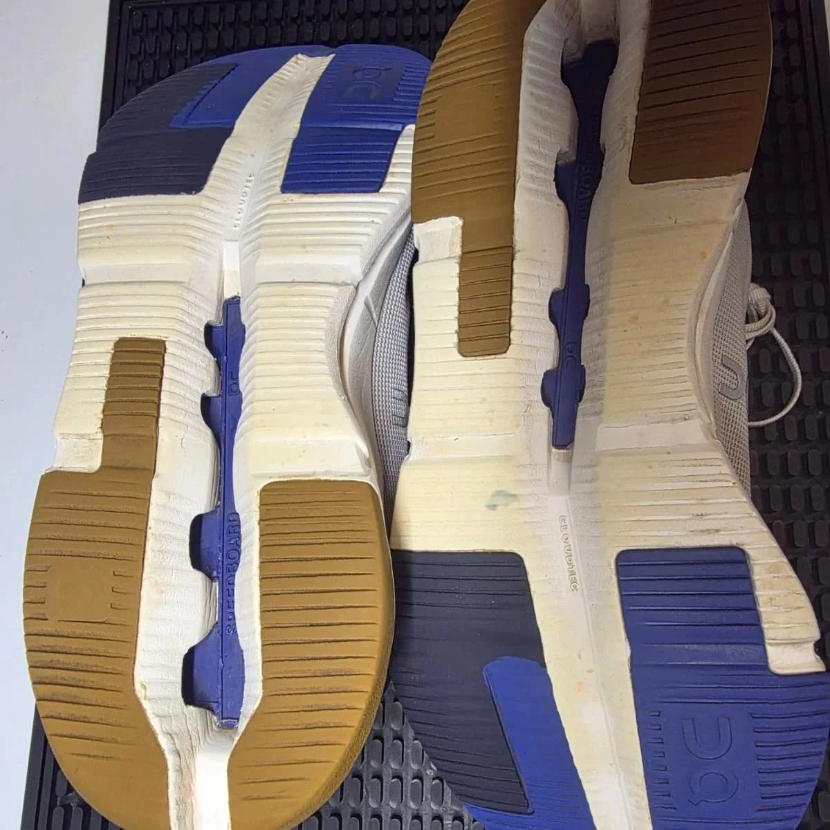 The image shows the soles of a pair of athletic sneakers with a color pattern of blue, black, beige, and white, placed on a black textured surface.
