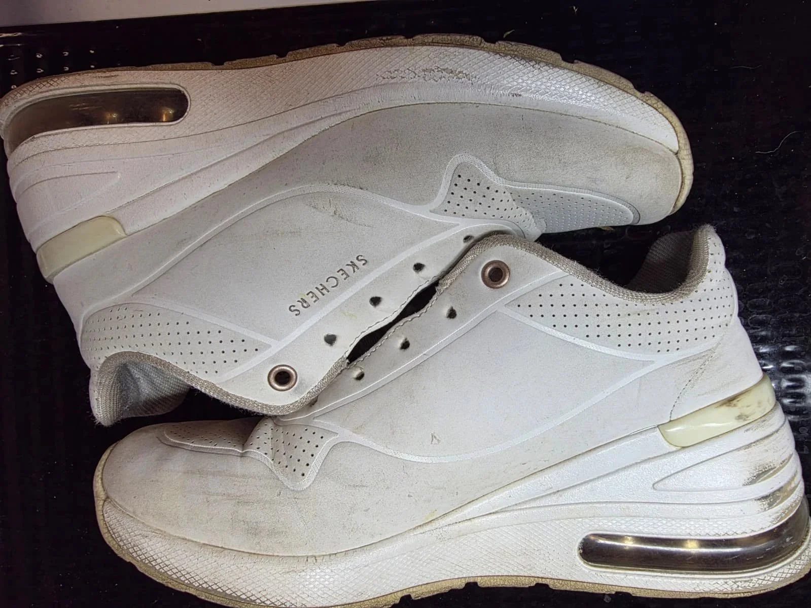 A pair of white Skechers sneakers with visible dirt and signs of wear, placed on a black textured surface.