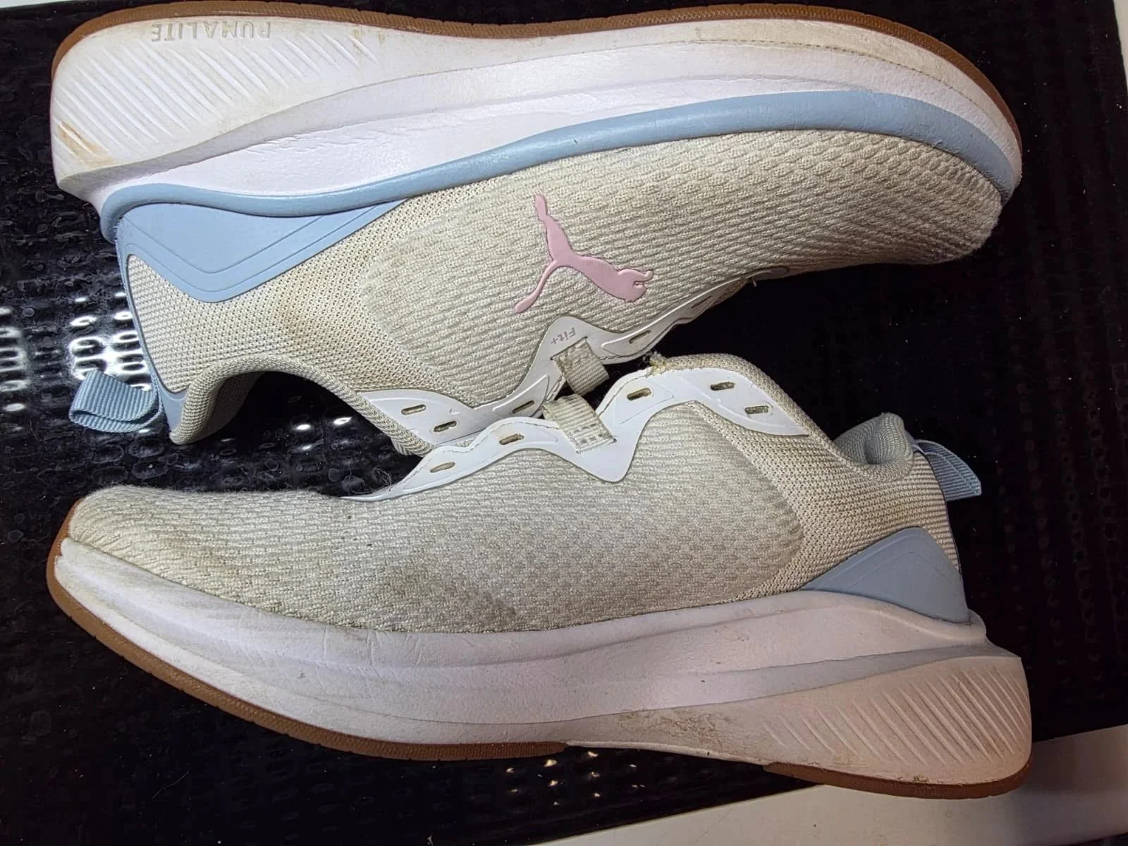 A pair of light-colored Puma sneakers with blue and black accents, placed on a black textured surface. The sneakers are new with tags attached.