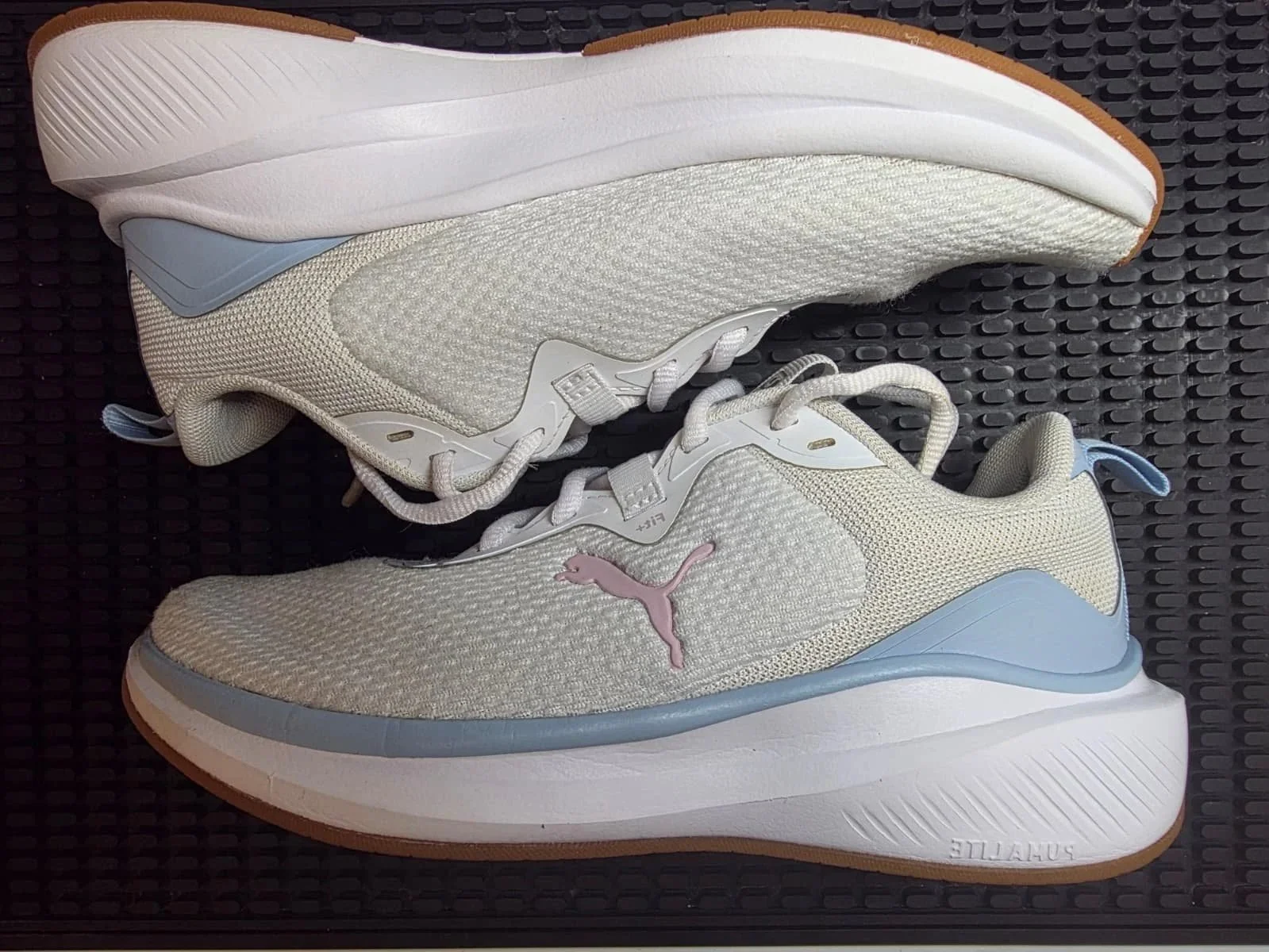 A pair of white Puma running shoes with pastel pink and blue accents, placed on a black textured surface.
