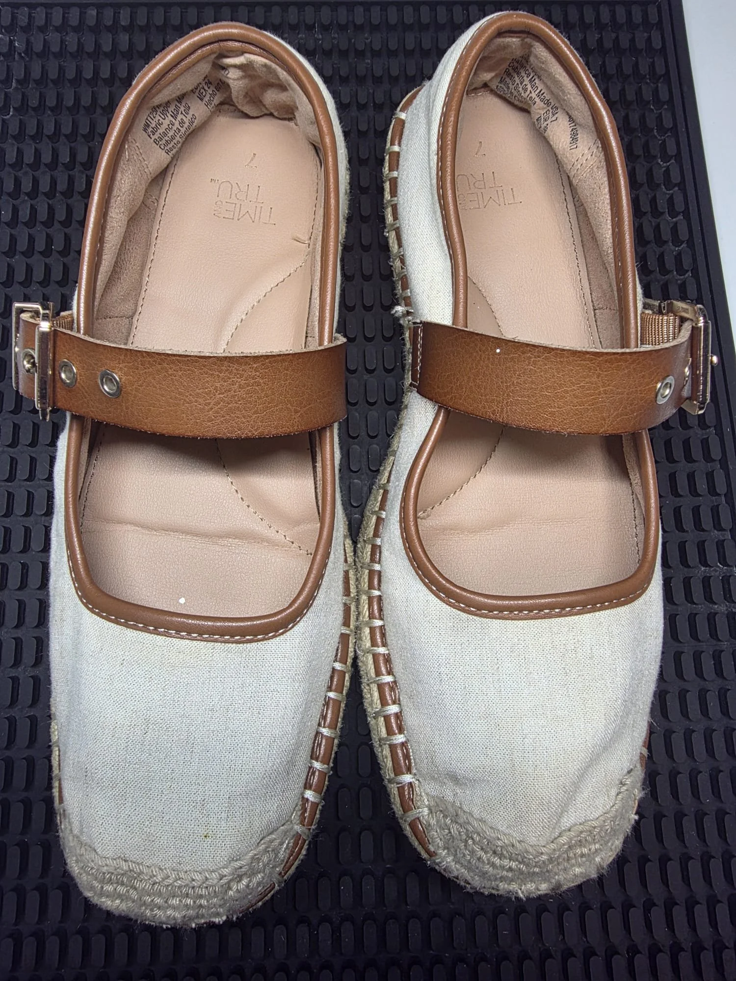 Pair of beige slip-on shoes with tan straps and stitching, placed on a black rubber mat.