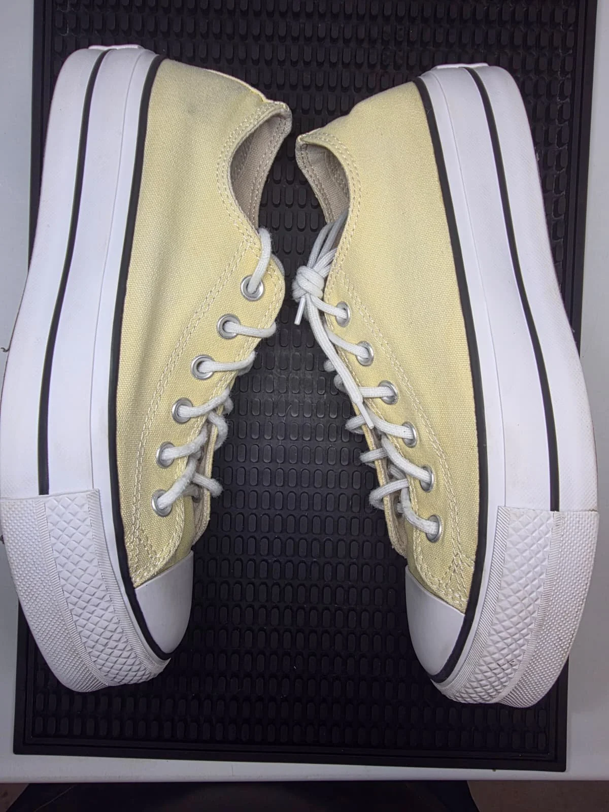 A pair of yellow canvas sneakers with white soles and white laces, placed on a black textured mat.