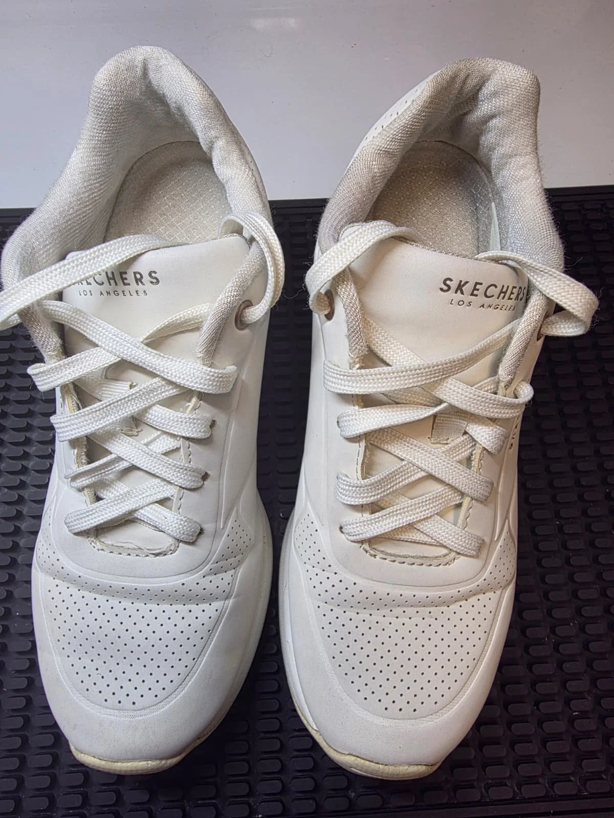 Pair of white Skechers sneakers with laces on black textured surface.
