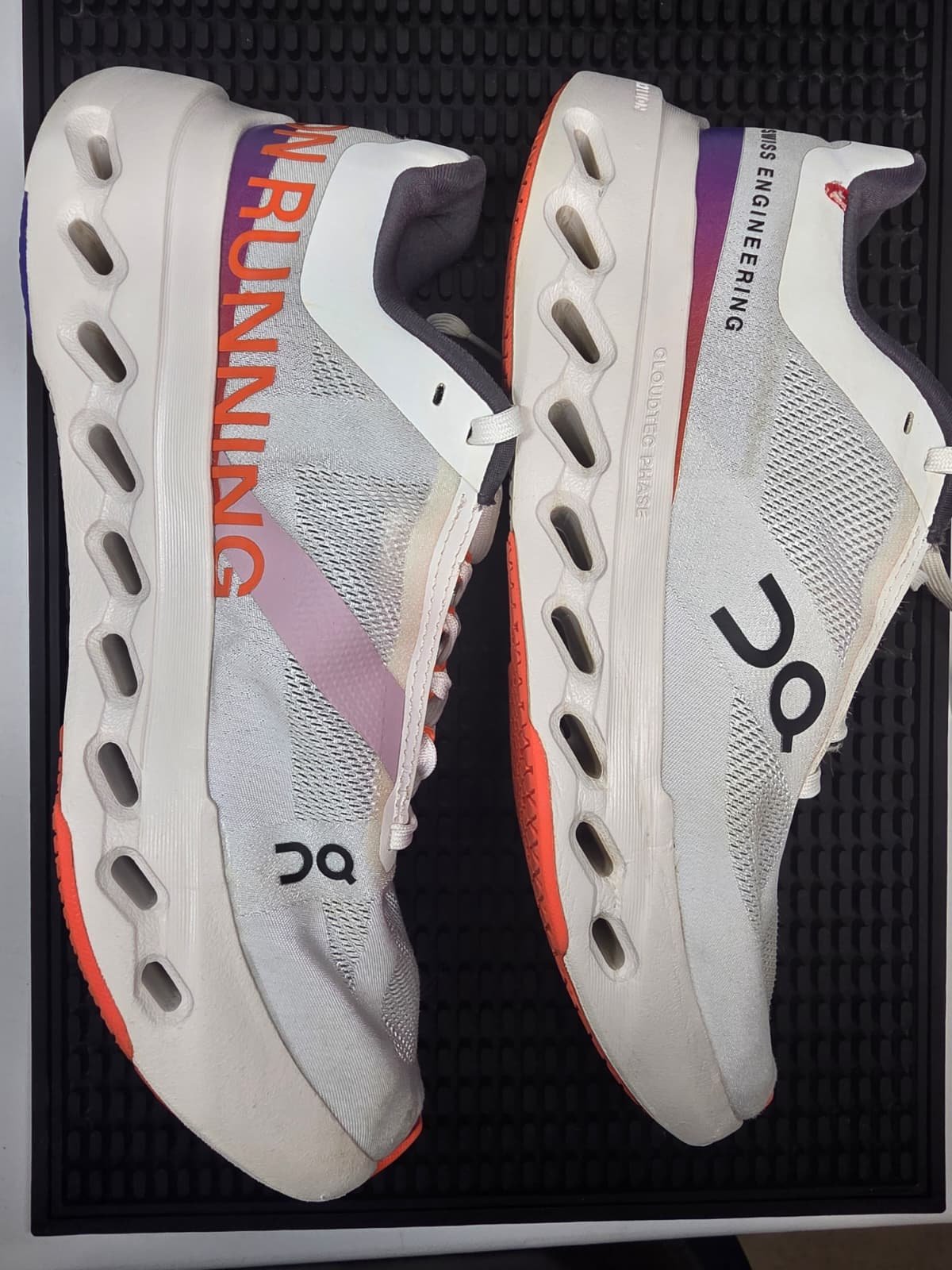 A pair of white running shoes with mesh fabric, branding, and an innovative sole design, placed on a black textured surface.