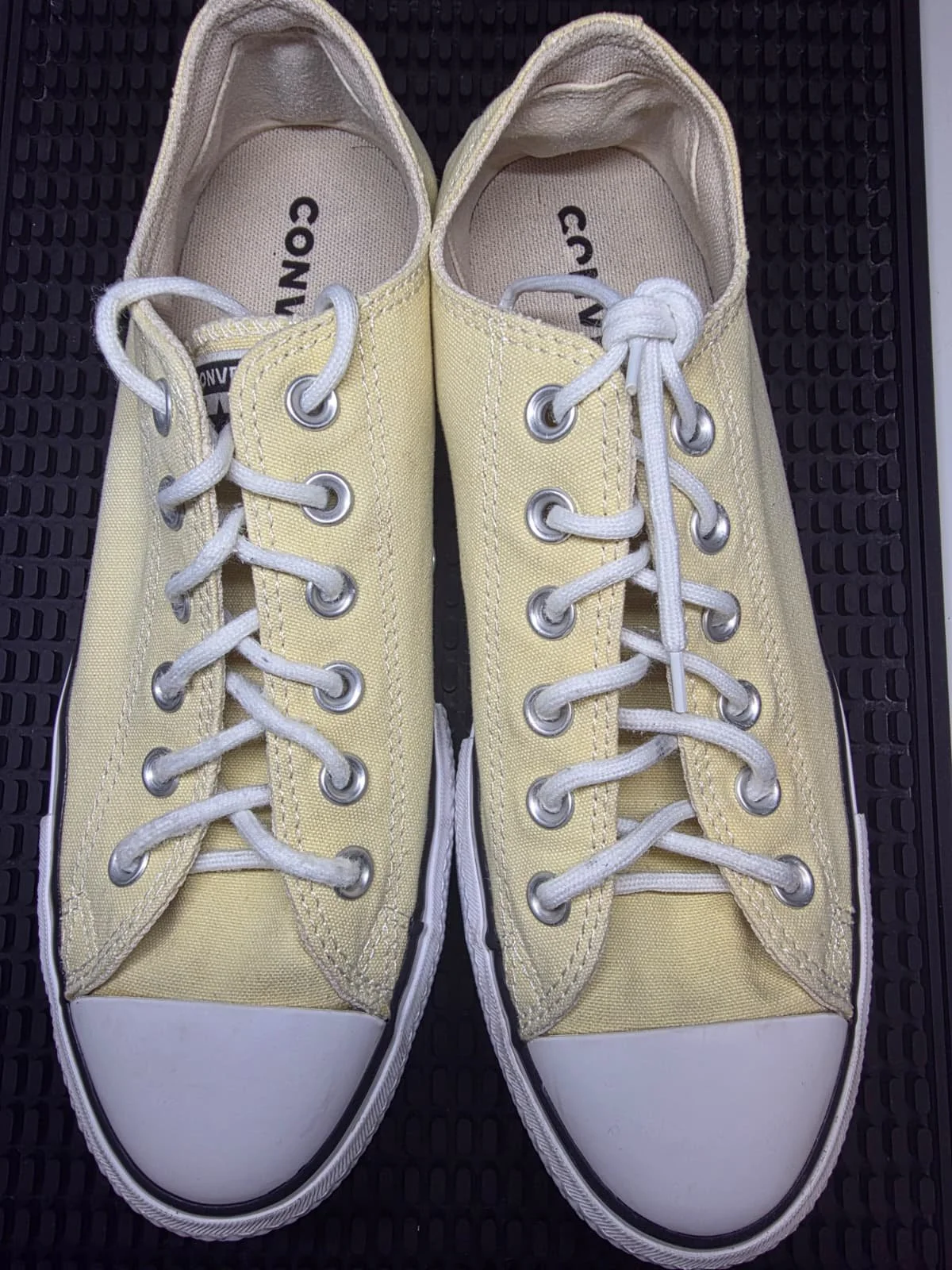 Pair of yellow Converse sneakers with white laces, white rubber toe caps, and beige insoles, placed on a black textured surface.