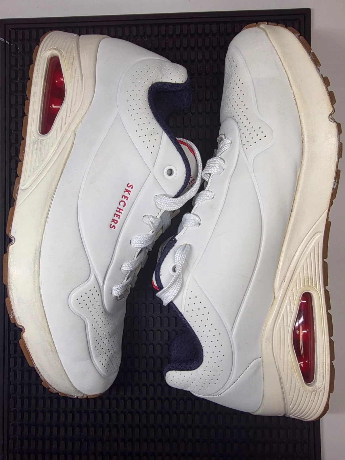 A pair of white Skechers sneakers with lace-up closure and red accents, placed on a black textured surface.