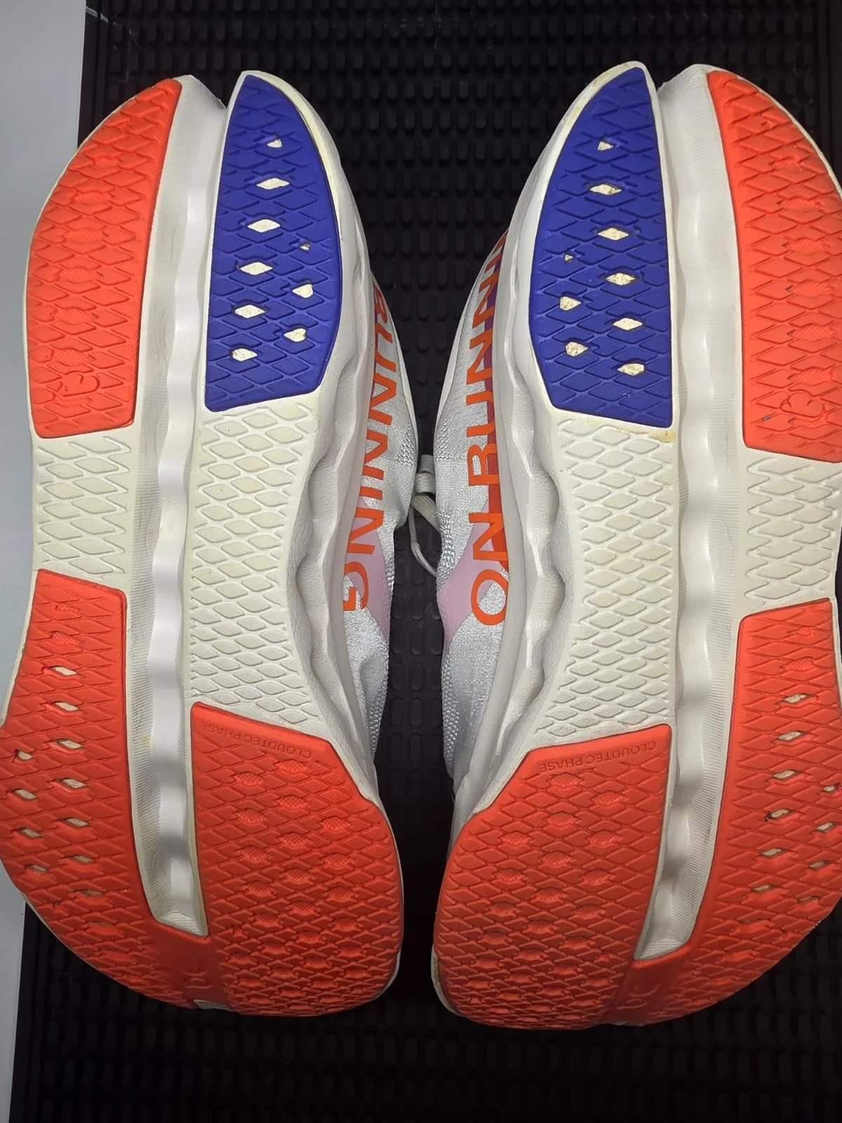 A pair of athletic shoes with orange, white, and blue soles, viewed from the bottom, placed on a black surface.