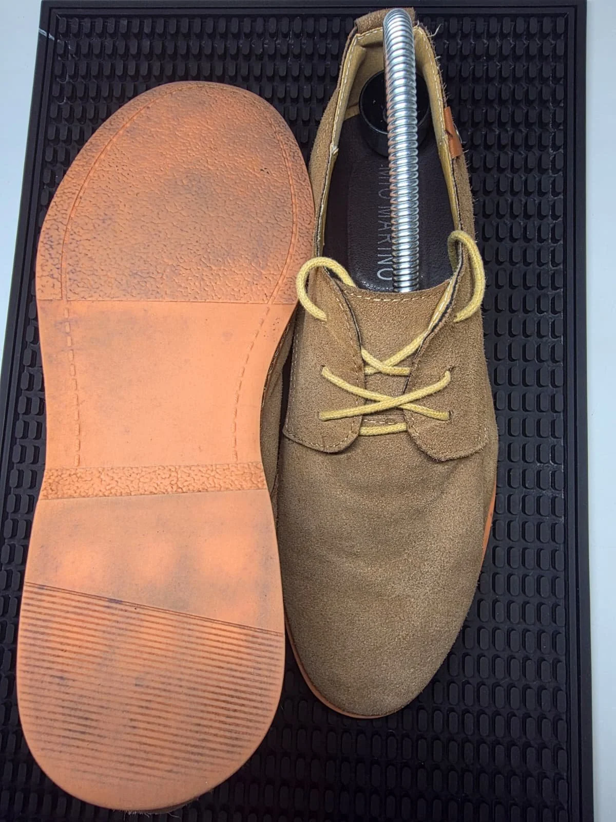A pair of brown suede shoes with yellow laces on a black rubber mat; one shoe is upright, showing the laces and interior, the other is lying on its side, showing the sole.