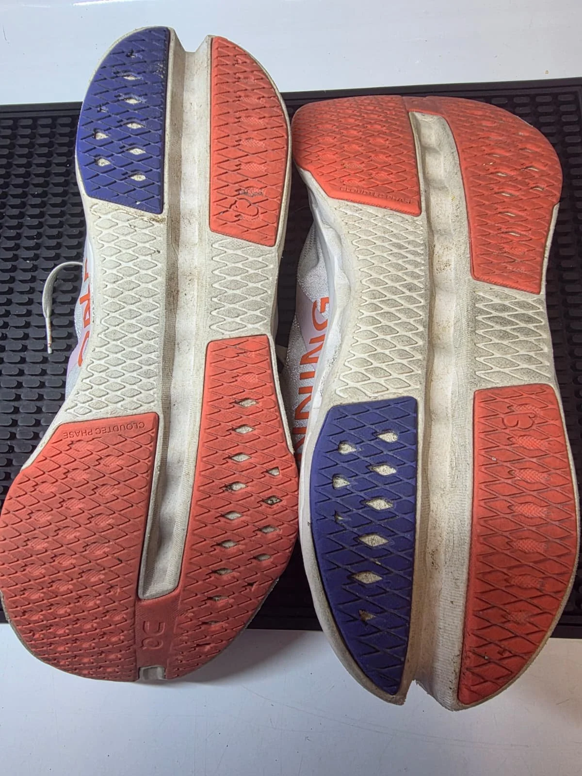 Worn running shoes with red and blue rubber soles, showing wear and dirt, placed soles-up on a black mat.