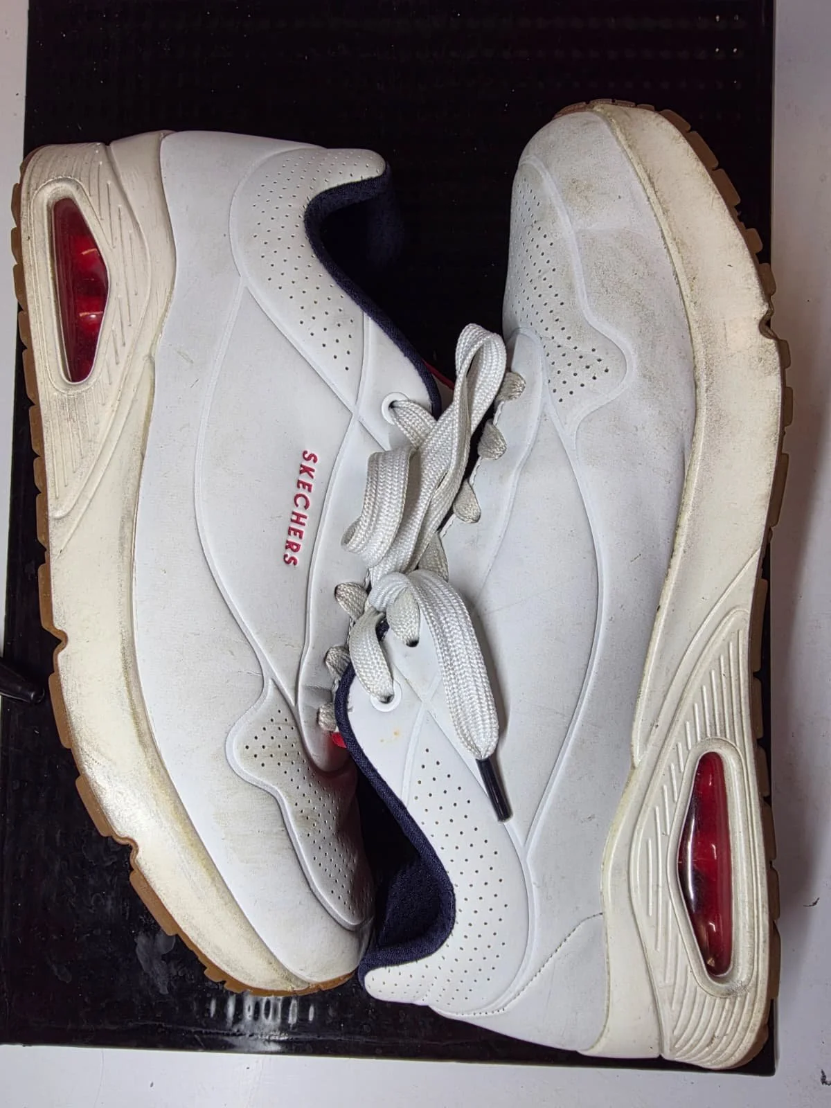 Pair of white Skechers sneakers with brown soles, positioned side by side with laces untied, placed on a black surface.