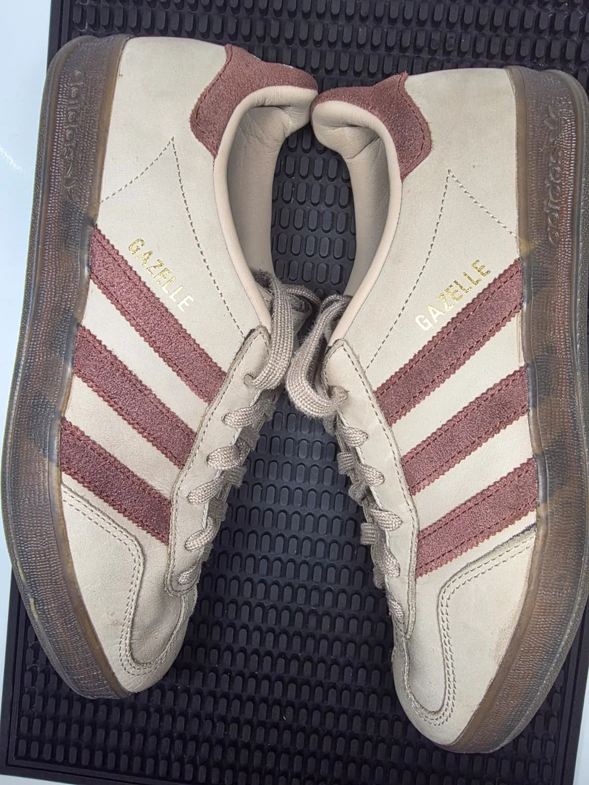 A pair of off-white Adidas Gazelle sneakers with pink suede stripes, placed on a black rubber mat.