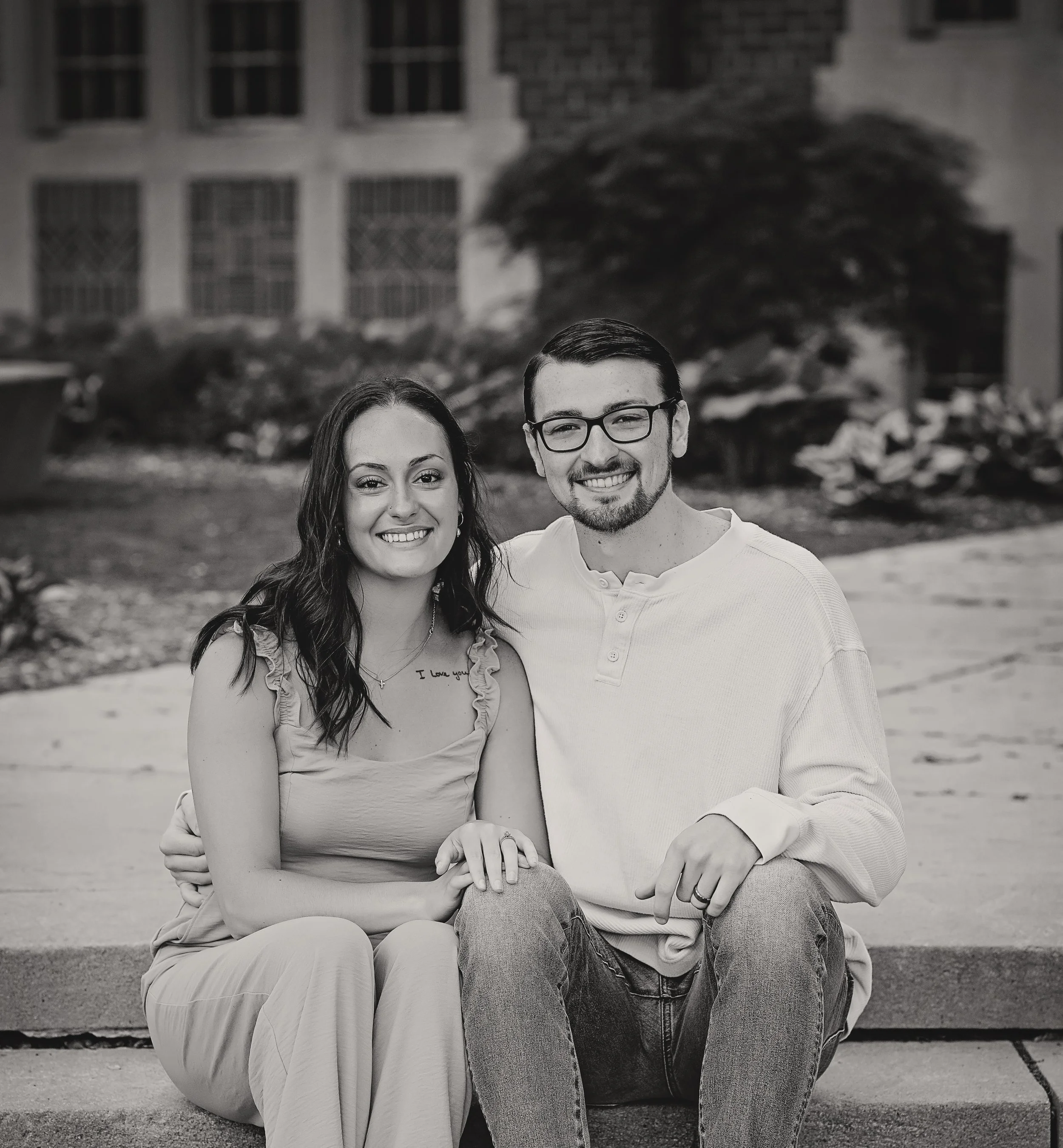 Black and white photograph of a smiling young woman with dark hair, wearing a light-colored top, and a young man with glasses, short dark hair, and a beard, wearing a light-colored long-sleeve shirt, sitting on a concrete bench outdoors with a building, trees, and rocks in the background.