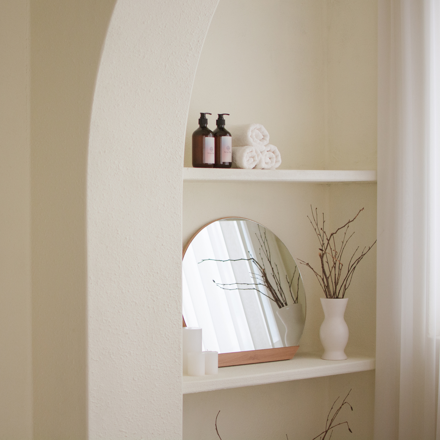 Decorative shelves with two brown bottles with black pump dispensers, white towels, white candles, a round mirror, and a white vase with dried branches on a light-colored wall.