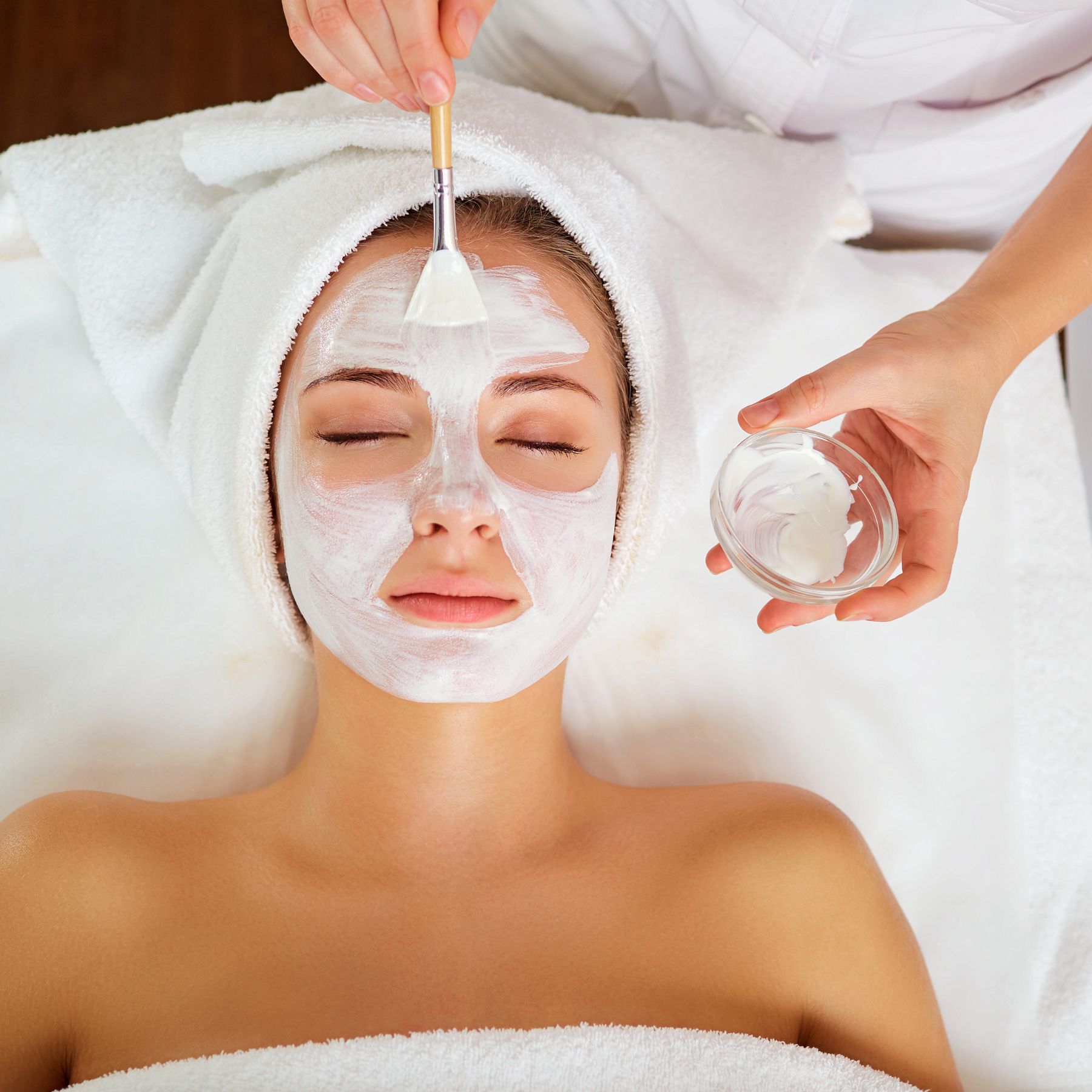 A woman lying with eyes closed, receiving a facial treatment. A person applies a white facial mask with a brush, holding a bowl of the mask, on the woman's face.