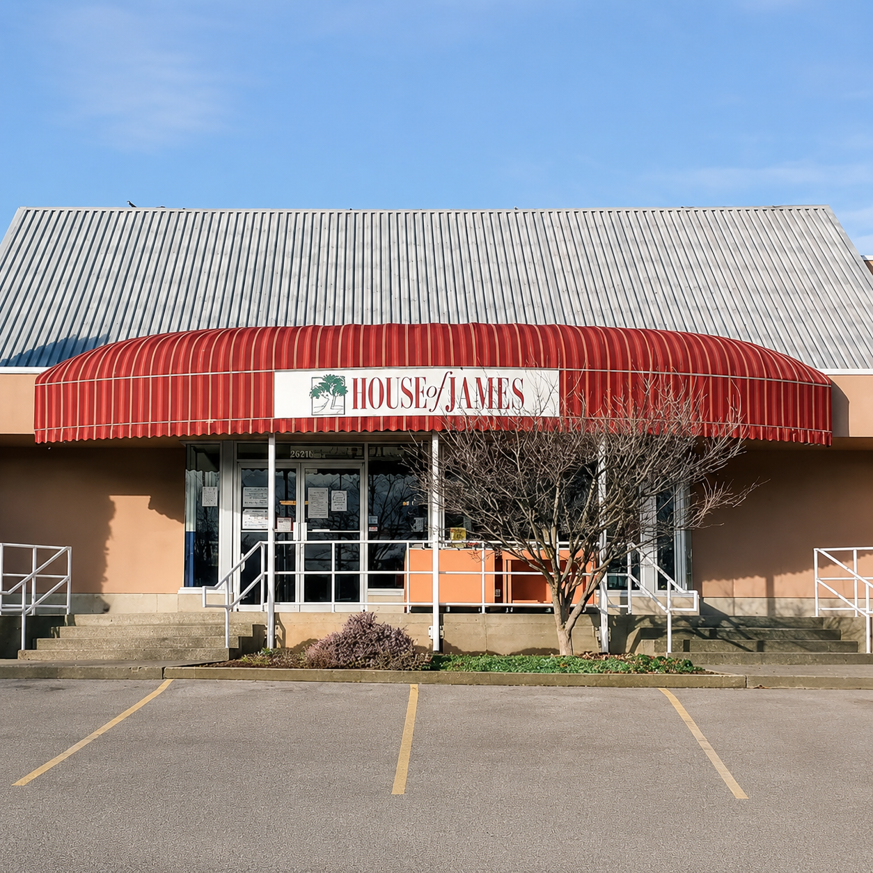 Exterior of a building with a red striped awning that reads 'House of James' and a leaf logo. The building has a metal roof and a small front porch with steps, surrounded by a parking lot with marked spaces and some bare trees.