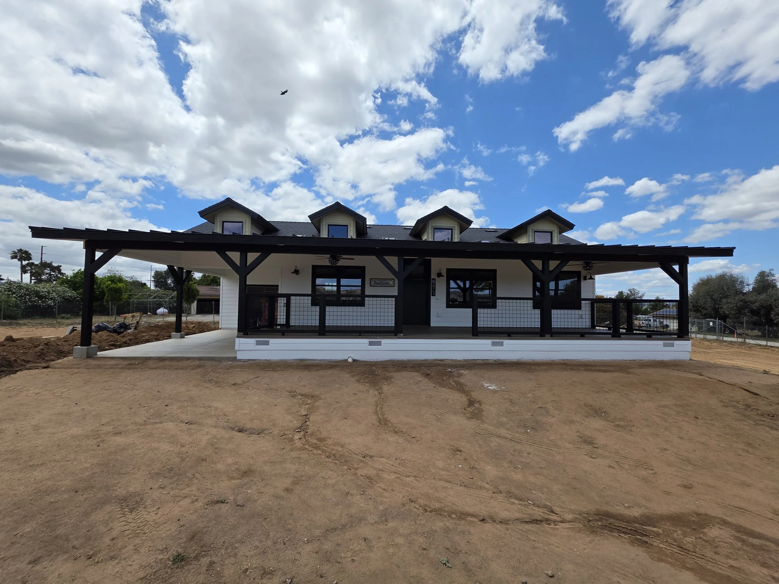 View of a house with a covered porch and dormer windows, under a partly cloudy sky, with dirt ground in the foreground.