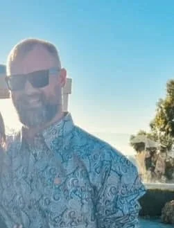A man with sunglasses and a beard wearing a patterned shirt outdoors with trees and a clear blue sky in the background.