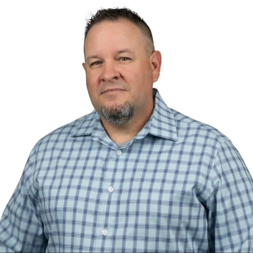 A man with short dark hair, a goatee, wearing a blue checkered button-up shirt, looking into the camera with a neutral expression.