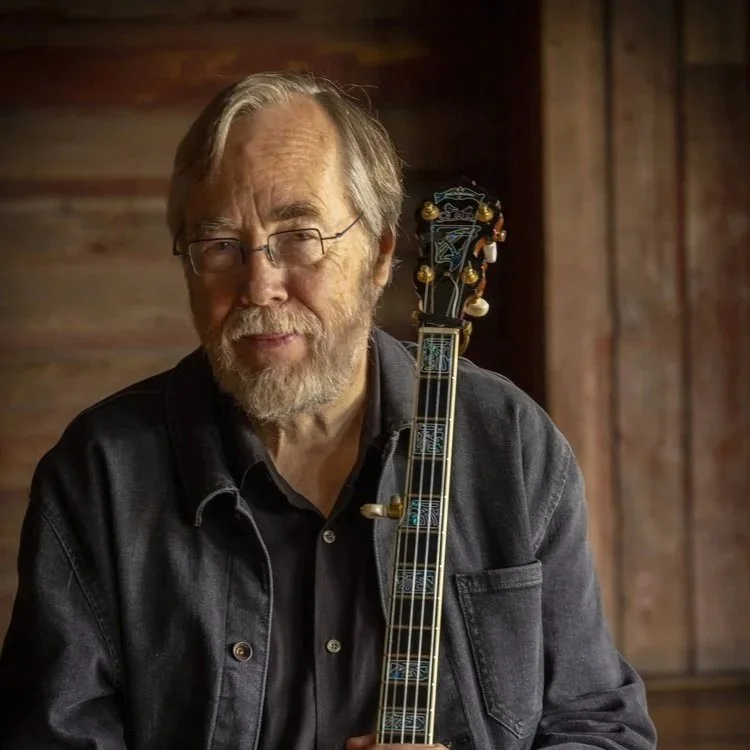 A bearded older man wearing glasses and a black denim jacket holding a decorative guitar, sitting against a wooden wall.