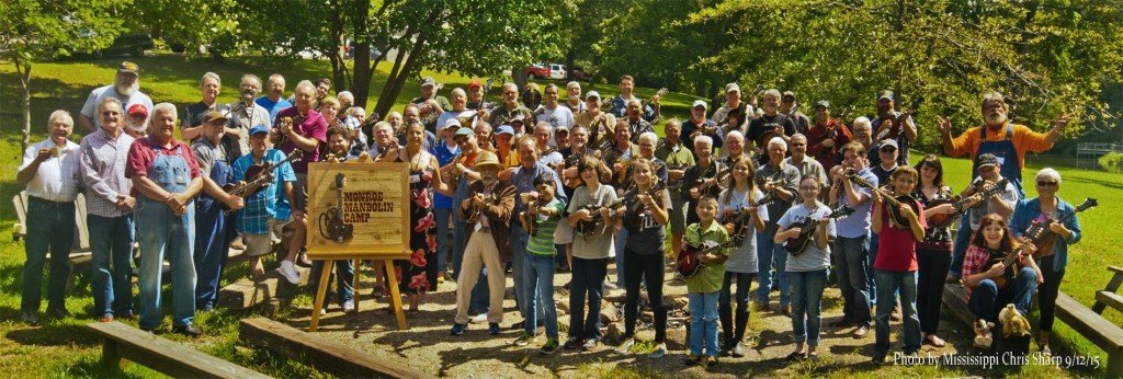 Group of children and adults playing guitars and posing outdoors during daytime at Monroe Maroon Camp, surrounded by trees.