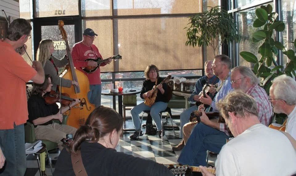 A group of people playing musical instruments together in a sunlit indoor space with large windows and plants.
