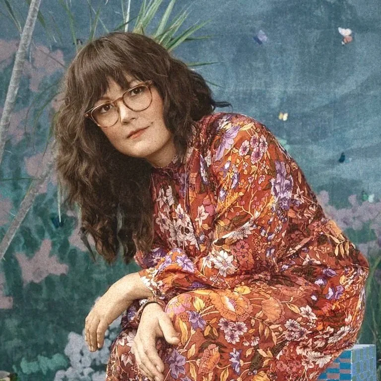 Young woman with wavy brown hair and glasses wearing a colorful floral dress, sitting outdoors near green plants and a painted background.