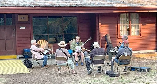 A group of seven elderly people sitting outdoors in front of a red building, with one woman playing a guitar.