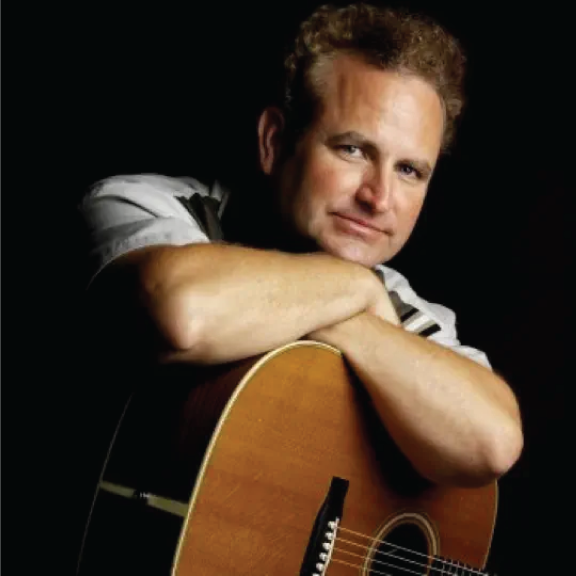 A man resting his head on his crossed arms, holding an acoustic guitar, with a serious expression, against a black background.