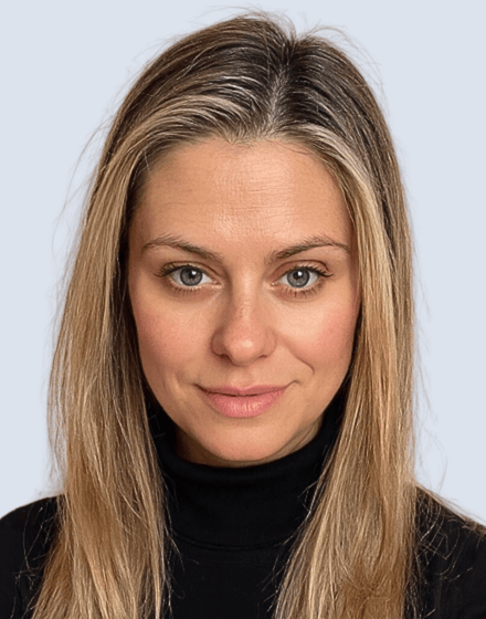 Close-up of a woman with blonde hair, blue eyes, wearing a black turtleneck, looking directly at the camera with a neutral expression.