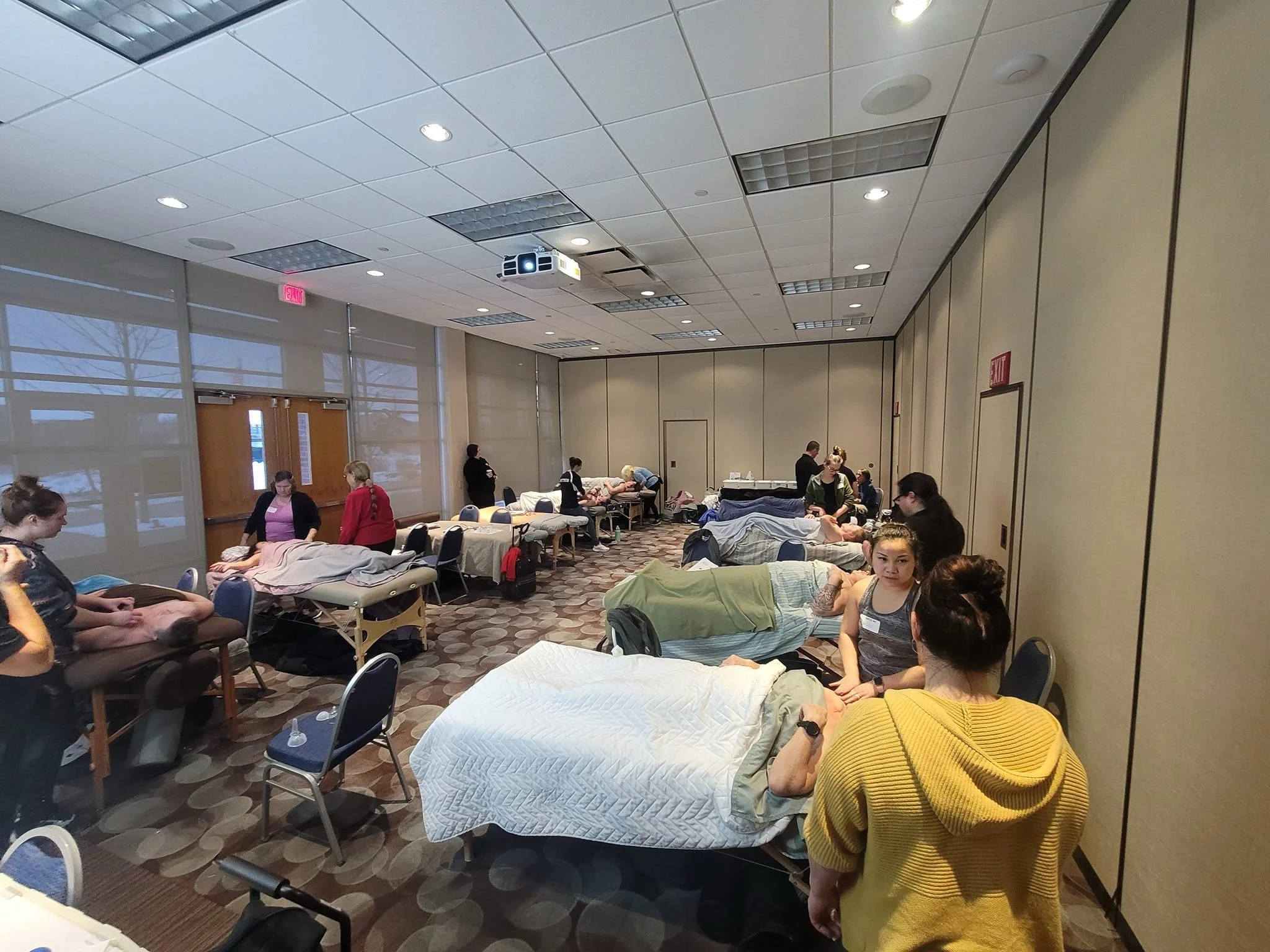 A room with people lying on beds and medical chairs, some being attended to by staff. The setting appears to be a blood donation or medical facility with a high ceiling, bright lighting, and large windows.