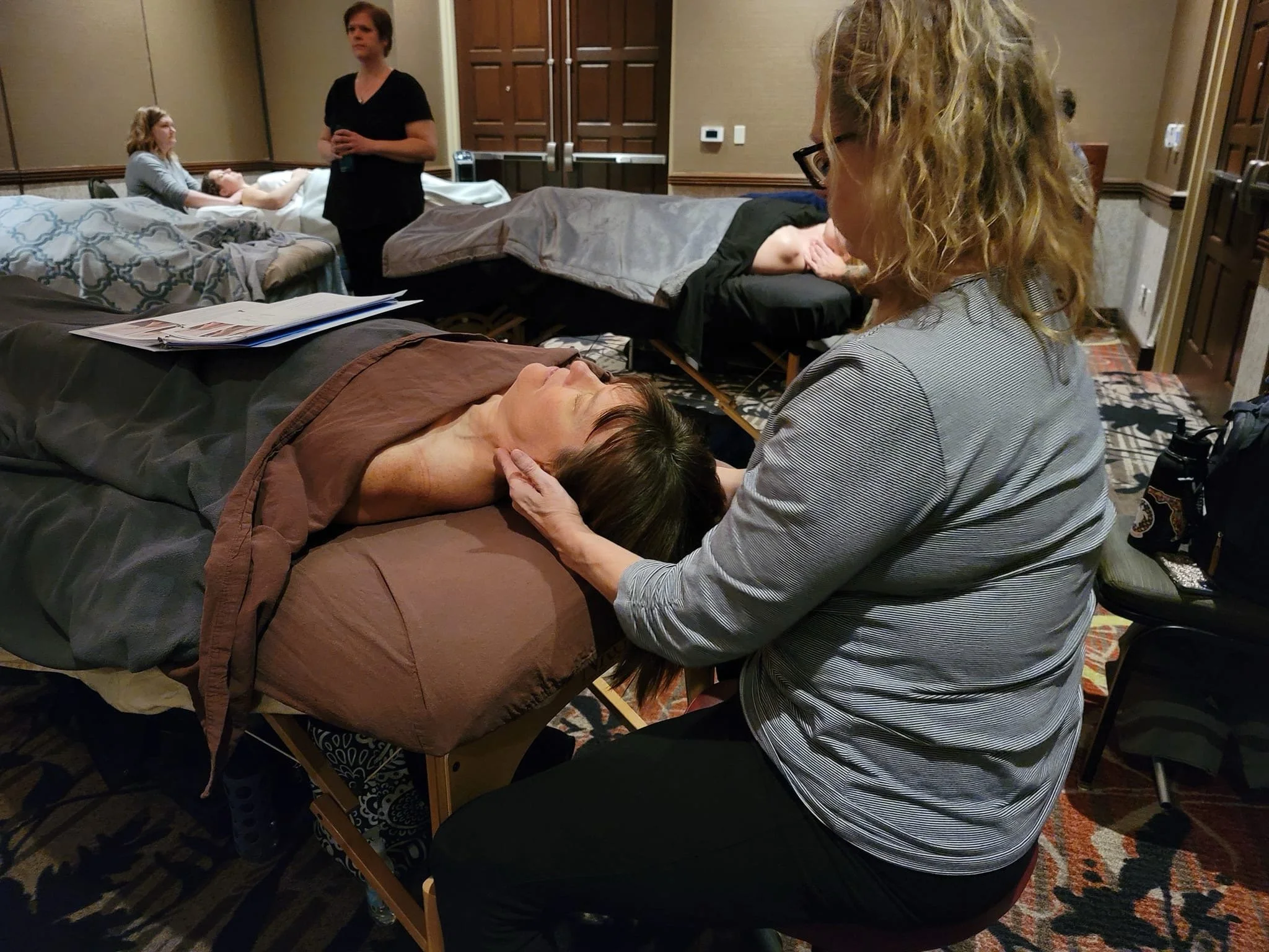 A woman receiving a massage in a spa or wellness center, lying on a massage table while a therapist is working on her neck and shoulders, with other clients and staff visible in the background.