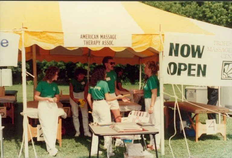 People working at a booth under a yellow and white tent, with a sign that says "NOW OPEN" and "AMERICAN MASSAGE THERAPY ASSOC." in green letters, outdoors on a grassy area.