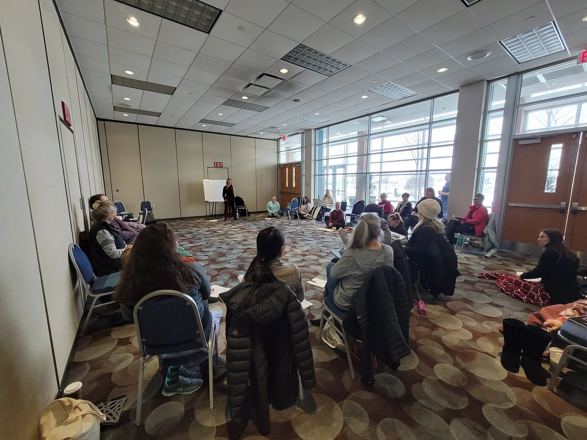 A group of people seated in chairs arranged in a circle inside a large meeting room with glass walls and ceiling lights, some appear to be participating in a discussion or workshop.