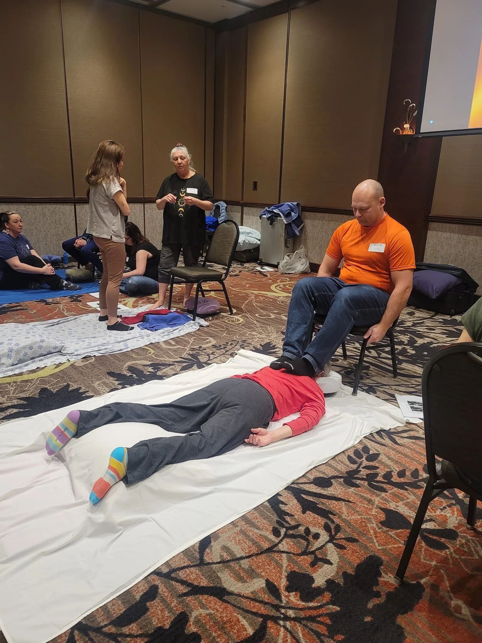 A group of people participating in a training or workshop session, with some sitting on the floor and others standing, in a conference room with a patterned carpet and a large screen.