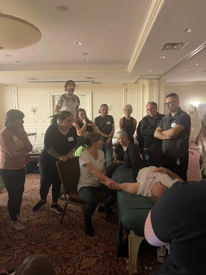 Group of people attending a CPR training session with a demonstration on a man lying on a bed.