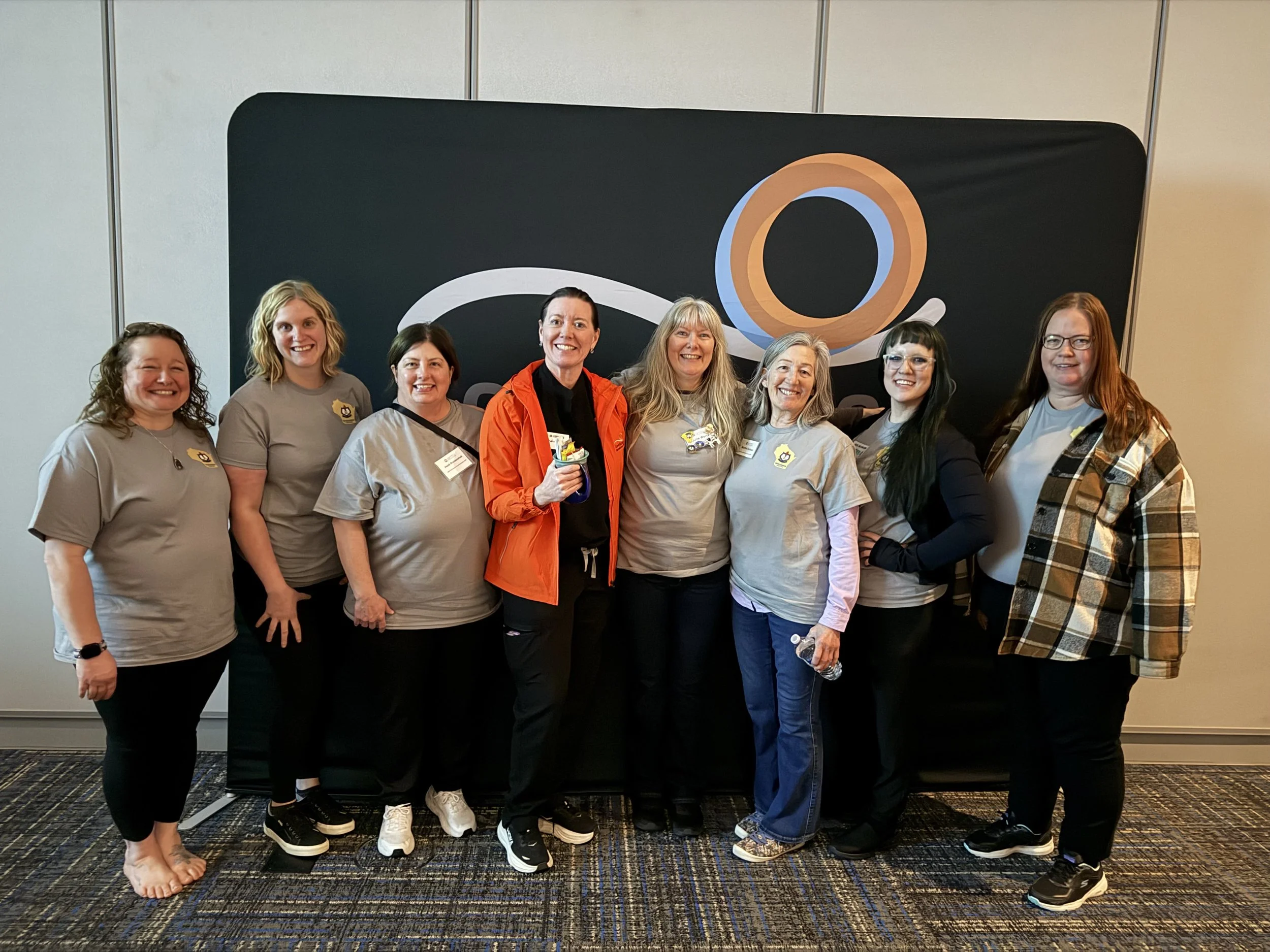 A group of nine women standing in front of a black backdrop with a logo, smiling for a photo at an indoor event.