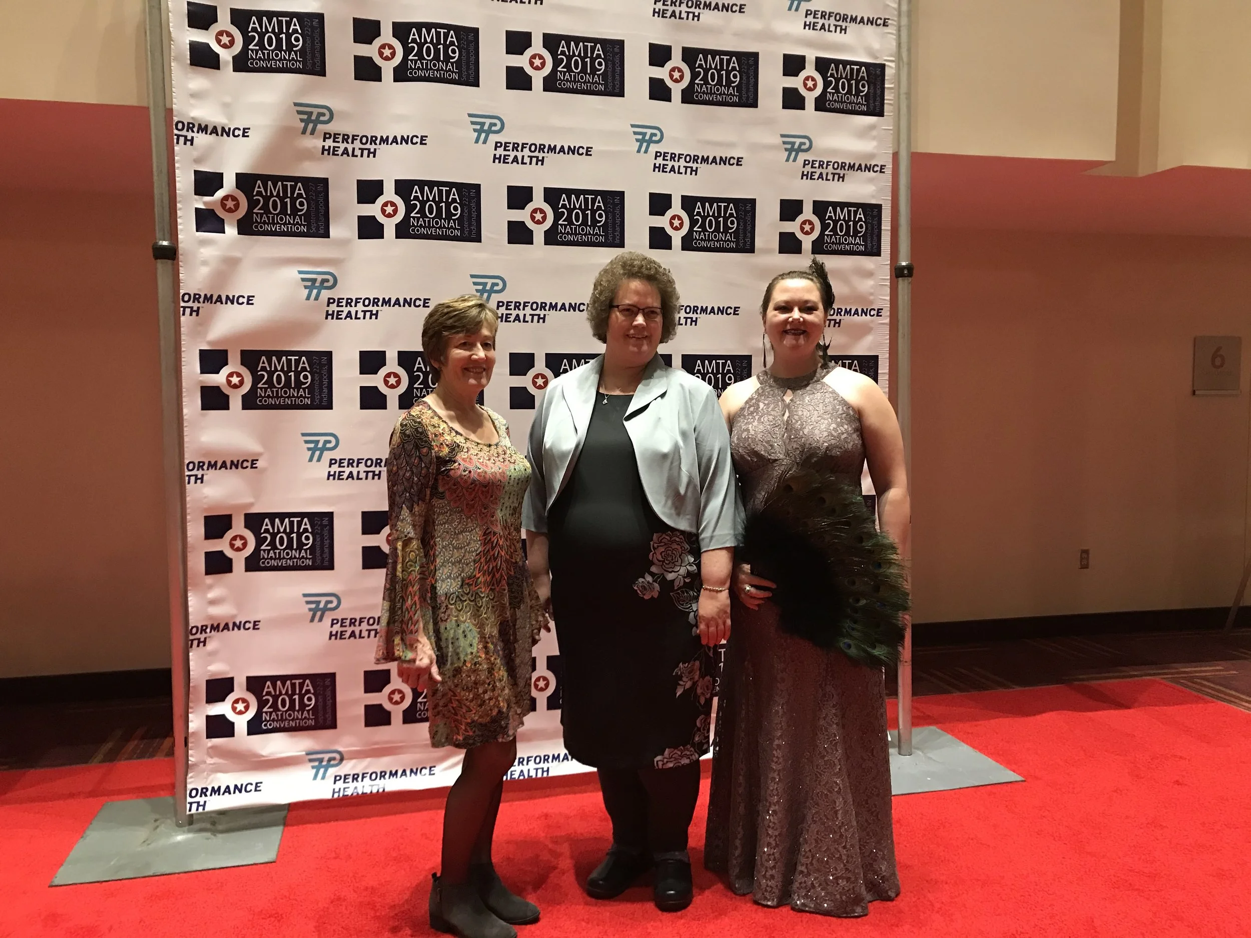 Three women standing together at the AMTA 2019 National Convention, posing for a photo in front of a backdrop with the event's logo. The backdrop features logos of Performance Health and the AMTA 2019 National Convention. They are all smiling and dre