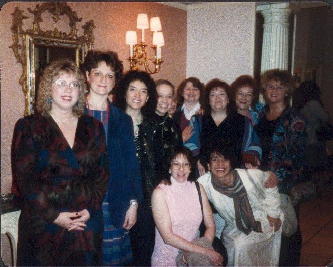 Group of women smiling and posing for a photo indoors, with a mirror and wall sconce in the background.