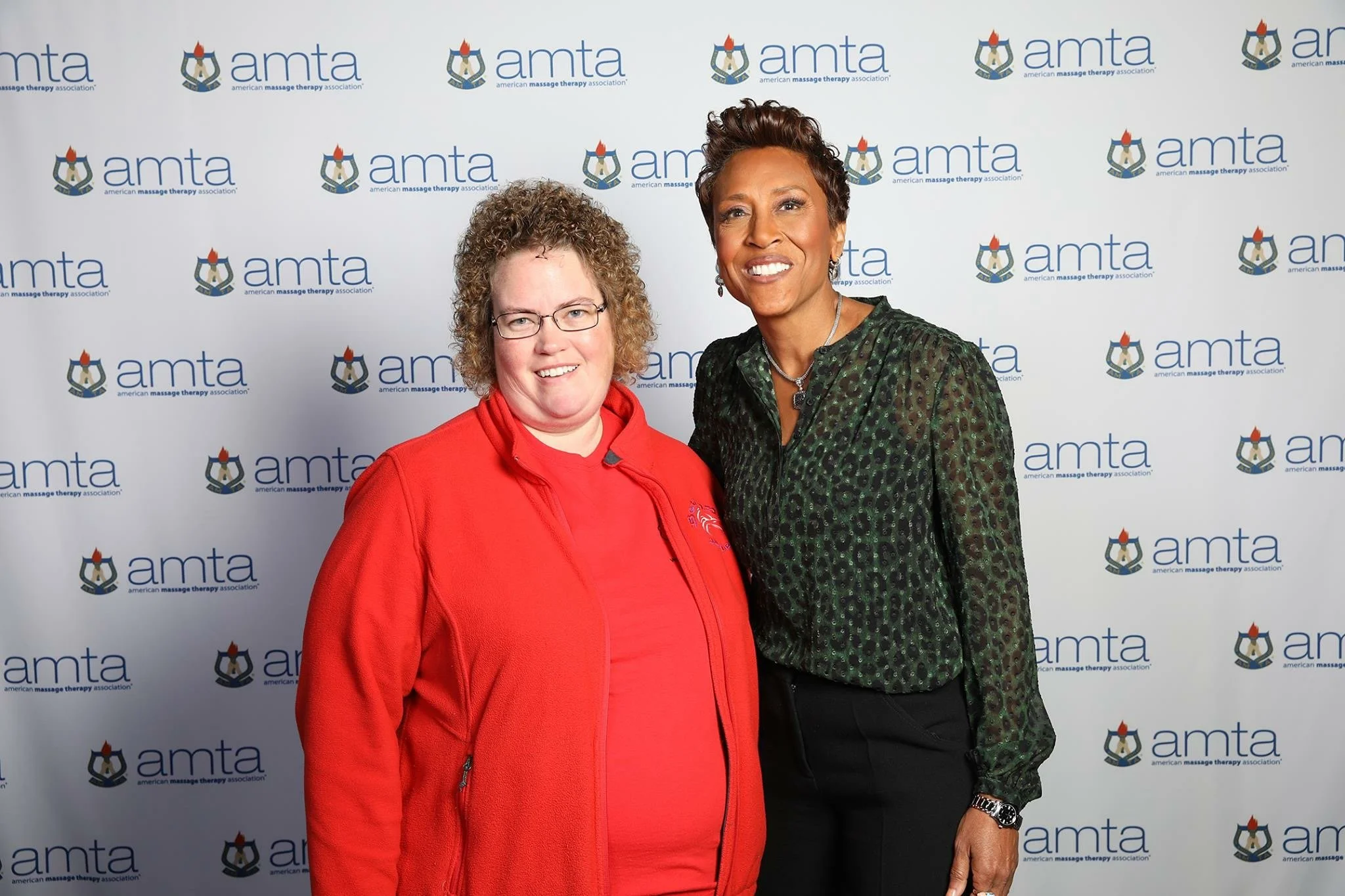Two women standing in front of a backdrop with the logo of the American Massage Therapy Association (AMTA). The woman on the left has curly blonde hair, glasses, and is wearing a red jacket. The woman on the right has short, styled brown hair and is 