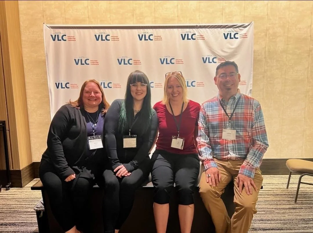 Group of four people sitting on a bench at a conference, smiling at the camera, with a banner behind them displaying the AMTA VLC logo.