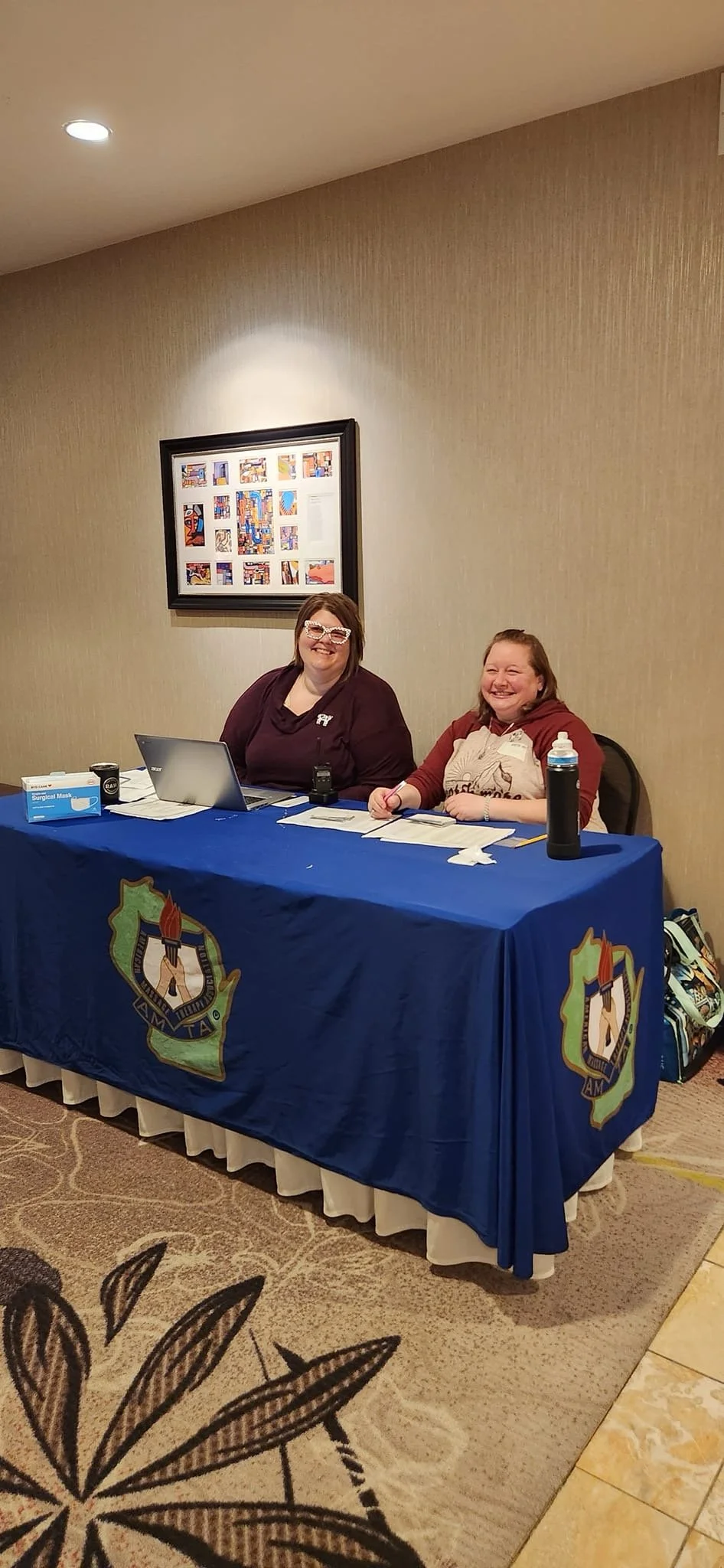 Two women sit at a table with a blue cloth featuring a badge logo, smiling at the camera. The table has papers, a laptop, a water bottle, and other items. The background has beige walls and framed artwork.