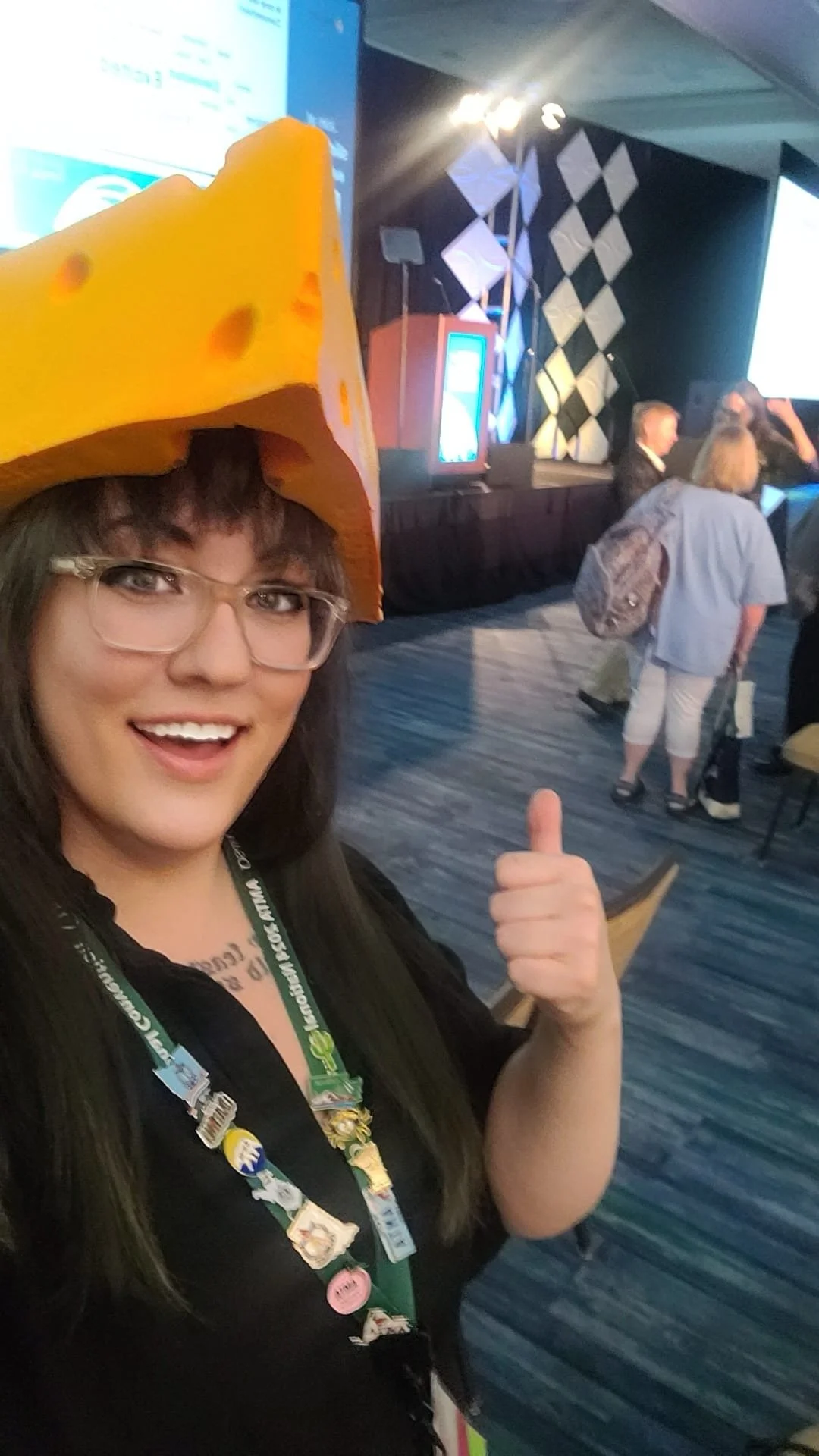 Woman at a conference wearing a cheese-shaped hat and a black shirt, with a conference badge and lanyard, smiling and giving a thumbs-up, in a room with a stage, people, and a large screen.