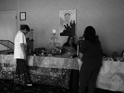 Two women and one man at an indoor craft or jewelry table, with various items displayed. The background has a framed artwork and a wall.