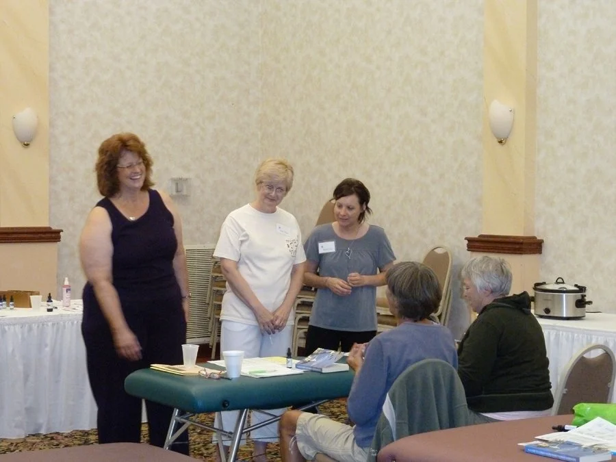Four women, two standing and two sitting, in a room with beige walls and a patterned carpet, engaged in a discussion or activity, possibly in a workshop or meeting setting.