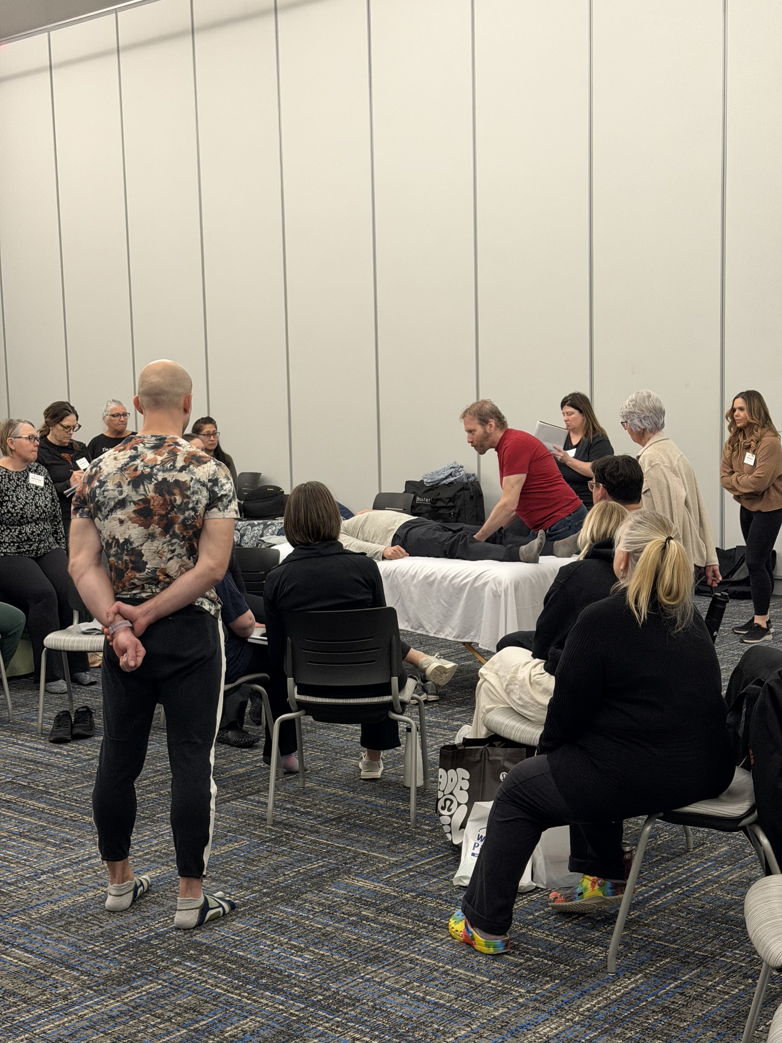 A group of people participating in a CPR training session in a conference room. Some are seated, some are standing, and a person is laying on a table for the demonstration. They are in a room with tall white walls and a striped carpet.