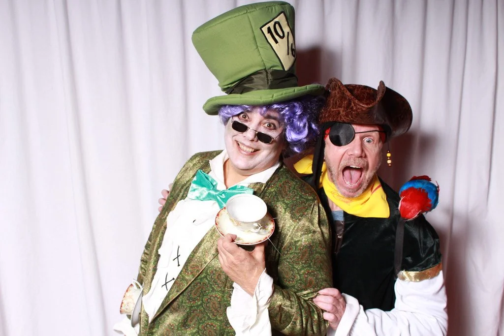 Two men dressed as characters from Alice in Wonderland, one as the Mad Hatter with a large green hat and purple wig, and the other as a pirate with a brown hat and eye patch, standing in front of a white curtain.