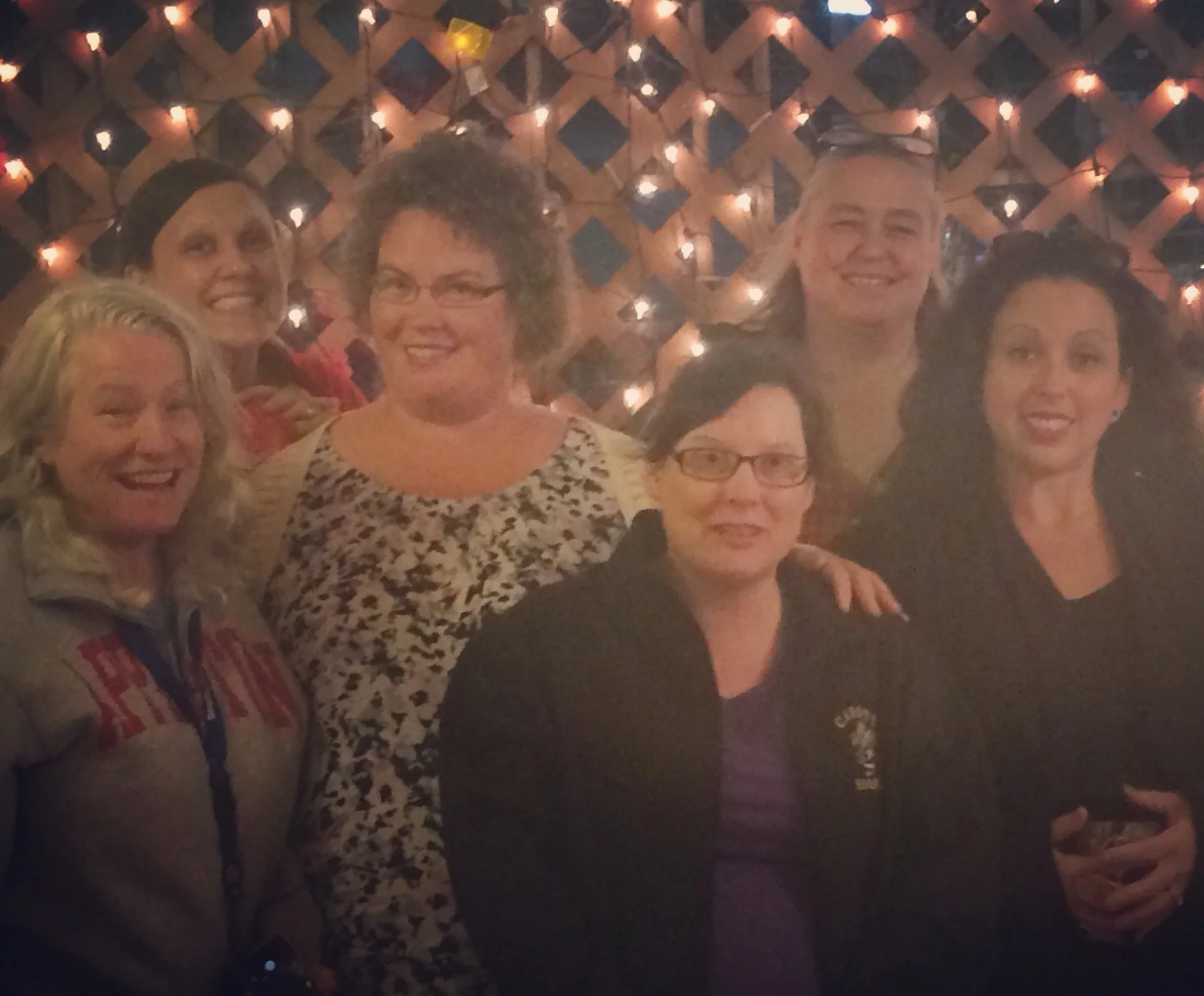 Group of six women smiling and posing for a photo in front of a wall decorated with string lights and black diamond-shaped panels.