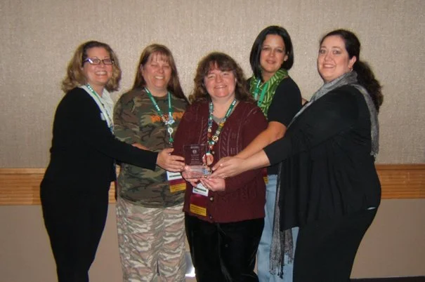 Five women standing together holding an award trophy, smiling at the camera in an indoor setting.