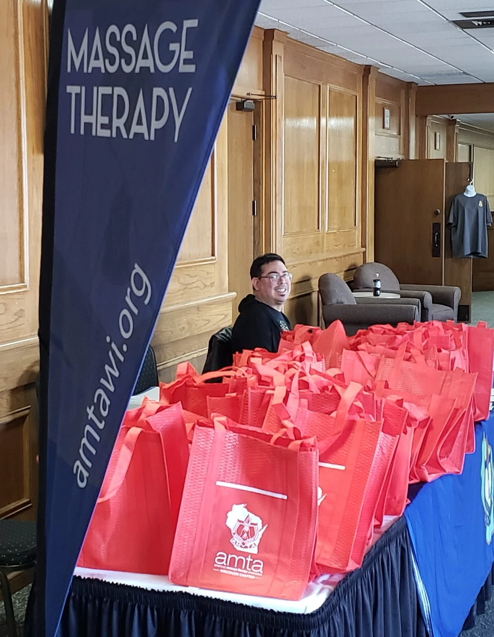 A table filled with red tote bags, a person sitting next to the bags, and a blue cloth banner with white text that reads 'Massage Therapy'.